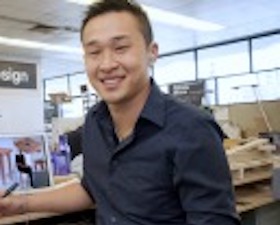 Young man, short dark hair, wearing dark shirt, holding pen, sitting at drawing table in design room, sign on wall says Industrial Design.