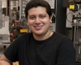 Young man, dark hair, in room with machining equipment.