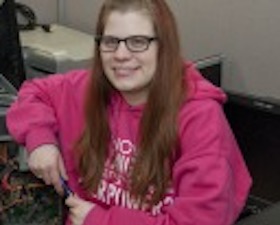 Young woman, long brown hair and glasses, pink hoodie, sitting by open computer showing inside circuit board.