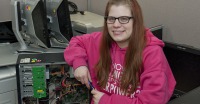 Young woman, long brown hair and glasses, pink hoodie, sitting by open computer showing inside circuit board.