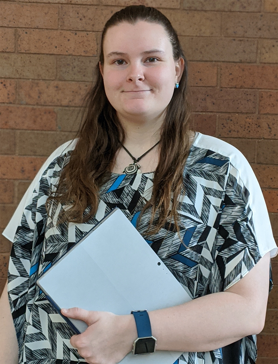 Photo of Kate Hawton, young woman with long brown hair over her shoulder smiling and holding notebook.