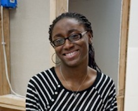 Young woman, dark skin, glasses, black and white diagonally striped top, black slacks, hands clasped together, sitting on desk.