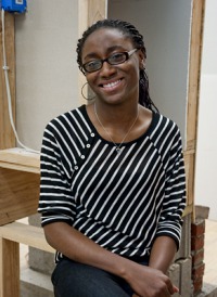 Young woman, dark skin, glasses, black and white diagonally striped top, black slacks, hands clasped together, sitting on desk.