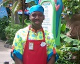 Young man, dark skin, wearing blue cap, multi-colored shirt and red apron, standing in front of tropical looking plants and building.