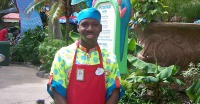 Young man, dark skin, wearing blue cap, multi-colored shirt and red apron, standing in front of tropical looking plants and building.