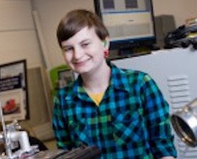 Young woman, short brown hair, wearing blue and black plaid shirt, standing in room of engineering equipment.