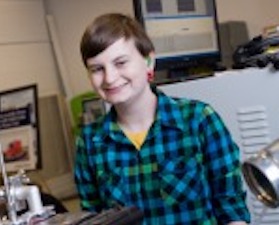 Young woman, short brown hair, wearing blue and black plaid shirt, standing in room of engineering equipment.