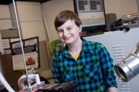 Young woman, short brown hair, wearing blue and black plaid shirt, standing in room of engineering equipment.