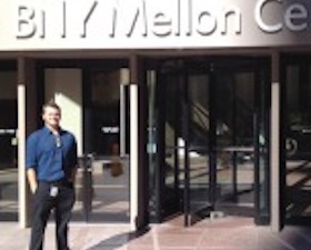 Young man, dark hair, hands in pockets, wearing dark shirt and slacks, standing in front of building that says BNY Mellon Center.