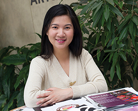 Young woman, dark hair, white v-neck top, sitting at table with graphic design materials.