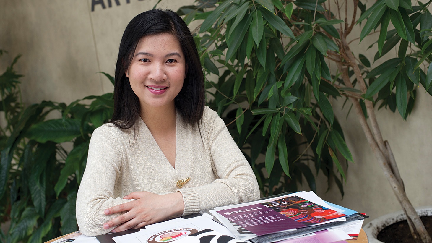 Young woman, dark hair, white v-neck top, sitting at table with graphic design materials.