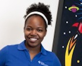 Young woman, dark skin, hair pulled back with white hair band, dark blue top, standing in front of poster with rocket and planets.