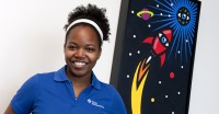 Young woman, dark skin, hair pulled back with white hair band, dark blue top, standing in front of poster with rocket and planets.