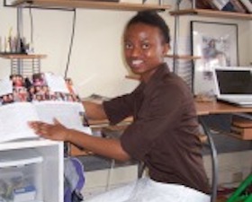 Young woman, dark skin, short dark hair, brown shirt, while slacks, sitting at office desk holding open book displaying photographs. 