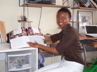 Young woman, dark skin, short dark hair, brown shirt, while slacks, sitting at office desk holding open book displaying photographs. 