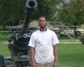 Young man, dark skin, short dark hair, white shirt and tan slacks, standing in field near military equipment and cannon.