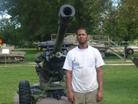 Young man, dark skin, short dark hair, white shirt and tan slacks, standing in field near military equipment and cannon.