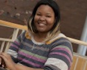 Young woman, dark skin, wearing striped sweater, sitting at table with laptop, stairway in background.