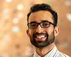 Photo of young man with dark hair and short beard, smiling, wearing dark glasses, striped shirt and patterned neck tie.