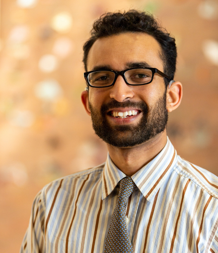 Photo of young man with dark hair and short beard, smiling, wearing dark glasses, striped shirt and patterned neck tie.