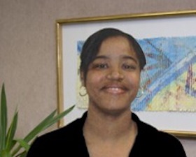 Young woman, dark skin, short dark hair, dark v-necked top, standing before potted plant and wall with framed art work.