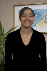 Young woman, dark skin, short dark hair, dark v-necked top, standing before potted plant and wall with framed art work.