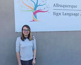 Photo of Malia Christenson, young woman with long dark blond hair, glasses, grey sweater dark pants and sneakers, standing in from of Sign Language Academy sign.