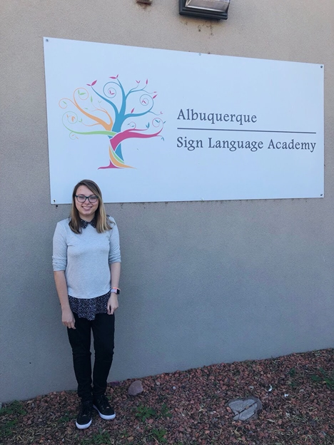 Photo of Malia Christenson, young woman with long dark blond hair, glasses, grey sweater dark pants and sneakers, standing in from of Sign Language Academy sign.