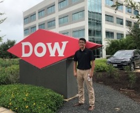 Young man, short hair, glasses, dark shirt, tan pants, outside standing in front of DOW sign.