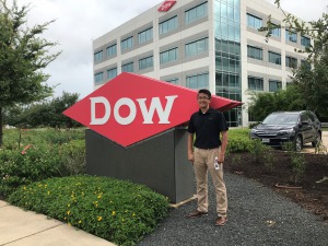 Young man, short hair, glasses, dark shirt, tan pants, outside standing in front of DOW sign.
