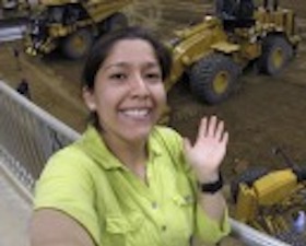 Young woman, short dark hair, wearing light green shirt, waving, standing on balcony over with tractor equipment below.