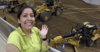 Young woman, short dark hair, wearing light green shirt, waving, standing on balcony over with tractor equipment below.
