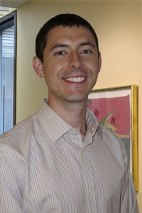 Young man, short dark hair, light colored striped shirt, standing in front of wall with framed artwork.