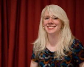 Young woman, with long blond hair, multi-colored top, sitting on chair with laptop open on lap, red curtain behind her.