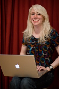 Young woman, with long blond hair, multi-colored top, sitting on chair with laptop open on lap, red curtain behind her.