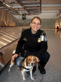 Young woman, brown hair tied back, wearing security officer uniform, kneeling next to beagle service dog.