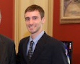 Young man, short dark hair, dressed in dark suit and tie, standing with an older man, in a government office.