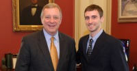 Young man, short dark hair, dressed in dark suit and tie, standing with an older man, in a government office.