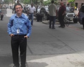 Young man, dark hair, glasses, blue shirt and dark slacks, hands in pockets, standing in front of building that says CBS Broadcasting.