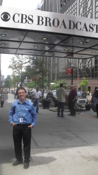 Young man, dark hair, glasses, blue shirt and dark slacks, hands in pockets, standing in front of building that says CBS Broadcasting.