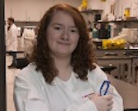 Young woman, dark hair, glasses in hand, arms crossed, wearing white lab coat in lab environment.