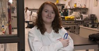 Young woman, dark hair, glasses in hand, arms crossed, wearing white lab coat in lab environment.