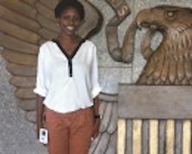 Young woman, dark skin, wearing white top and brown pants, standing in front of wooden carving of eagle, words US.