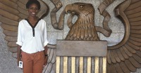 Young woman, dark skin, wearing white top and brown pants, standing in front of wooden carving of eagle, words US.