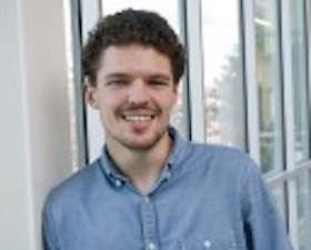 Young man, short dark hair, short mustache and beard, blue shirt, leaning on wall of windows.