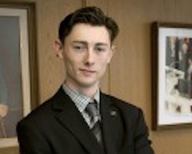 Young man, short dark hair, wearing dark suit and tie, standing in front of wall with two framed pictures.