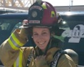 Young woman in firefighter gear and helmet standing in front of vehicle with word "Chief 2" on window.