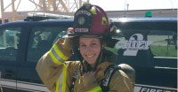 Young woman in firefighter gear and helmet standing in front of vehicle with word "Chief 2" on window.