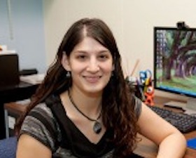 Young woman, long dark hair, wearing striped v-neck top, necklace, elbow up on desk, computer monitor behind her displays colorful trees.