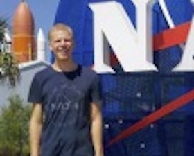 Young man, blond hair, wearing navy blue t-shirt with words NASA, standing in front of NASA statue.